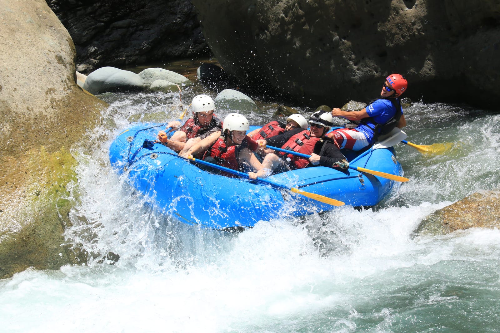 A group in helmets and life vests paddling a blue raft through whitewater rapids