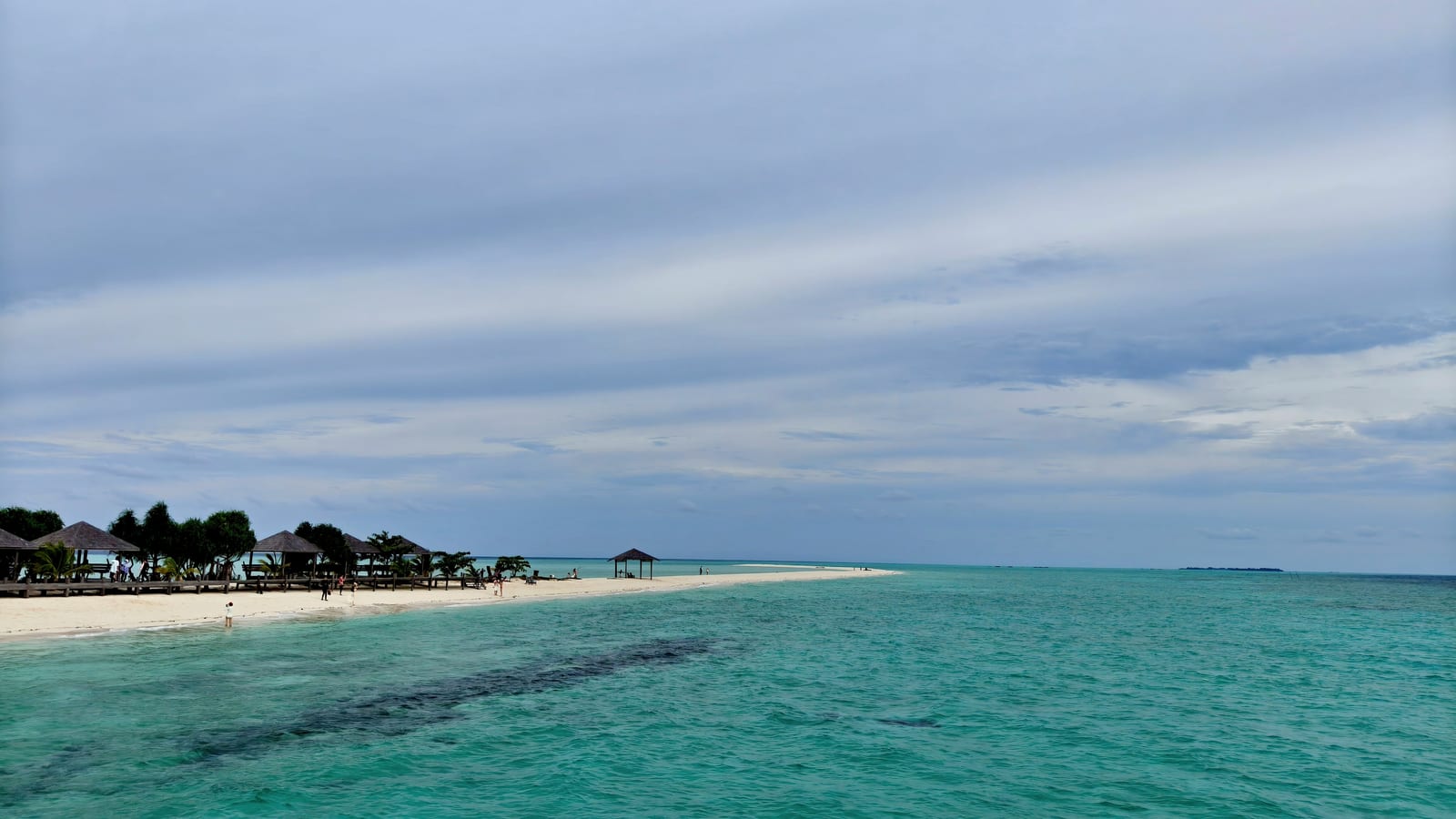 A low sand spit reaching into calm turquoise water under an overcast sky, with thatched shelters along it