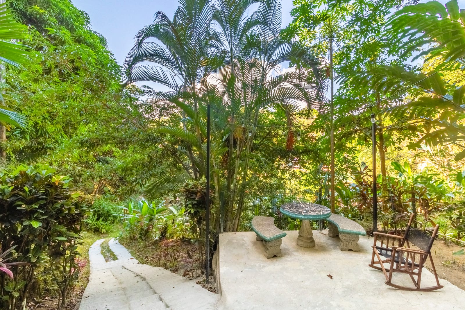 A round stone table with matching benches on a paved clearing, under tall palms and jungle planting