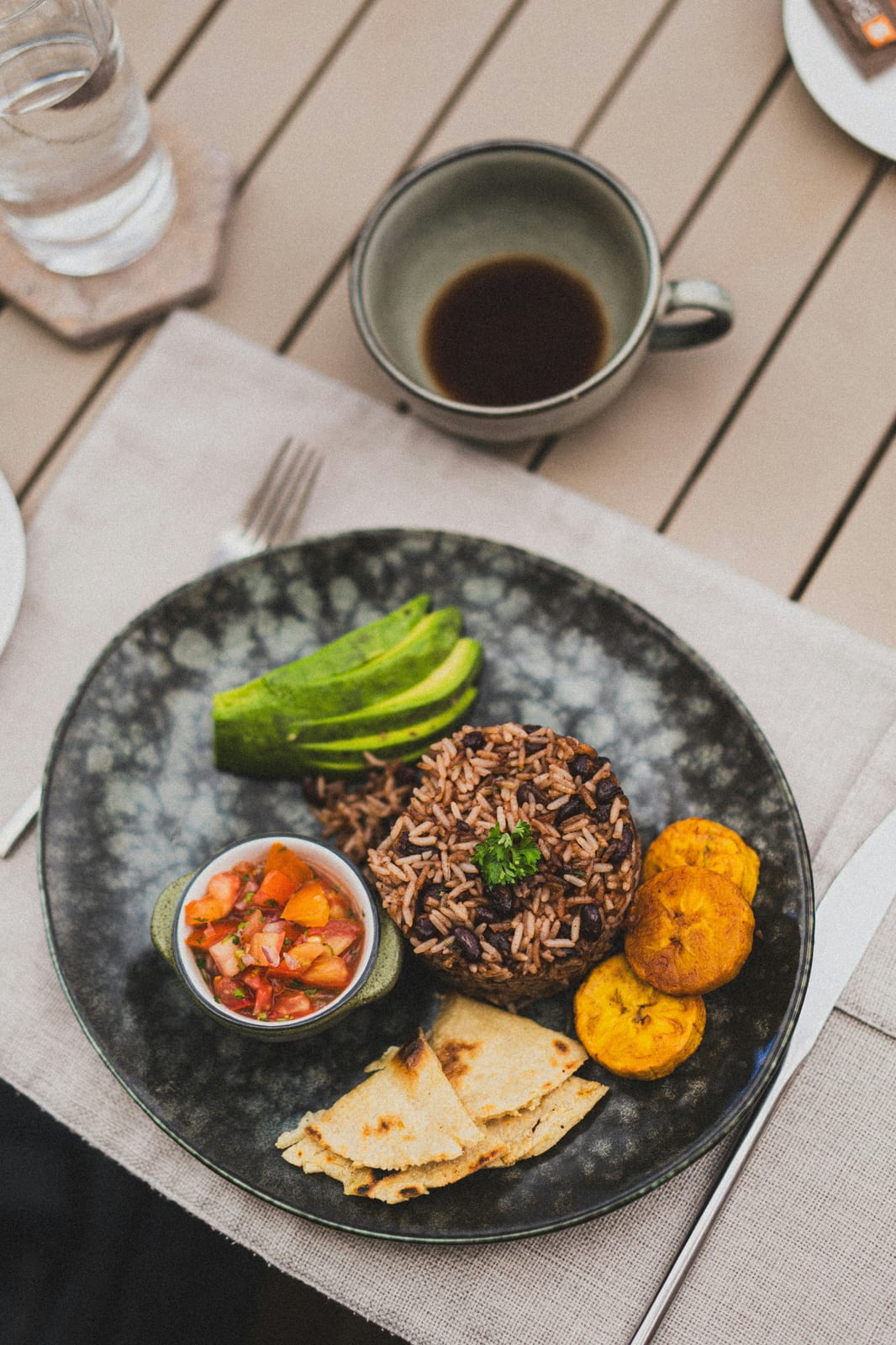 A Costa Rican plate of gallo pinto with avocado, pico de gallo, fried plantain and tortillas, beside a cup of black coffee