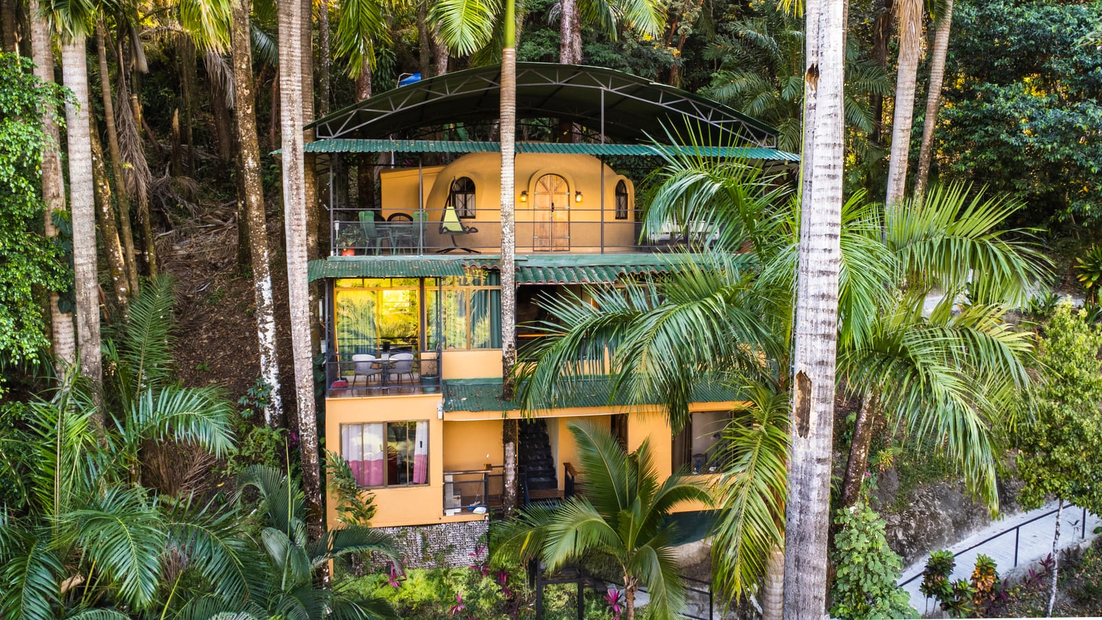 A two storey guest building in yellow with green metal roofs and arched balconies, seen from the air between tall palm trunks on a steep hillside
