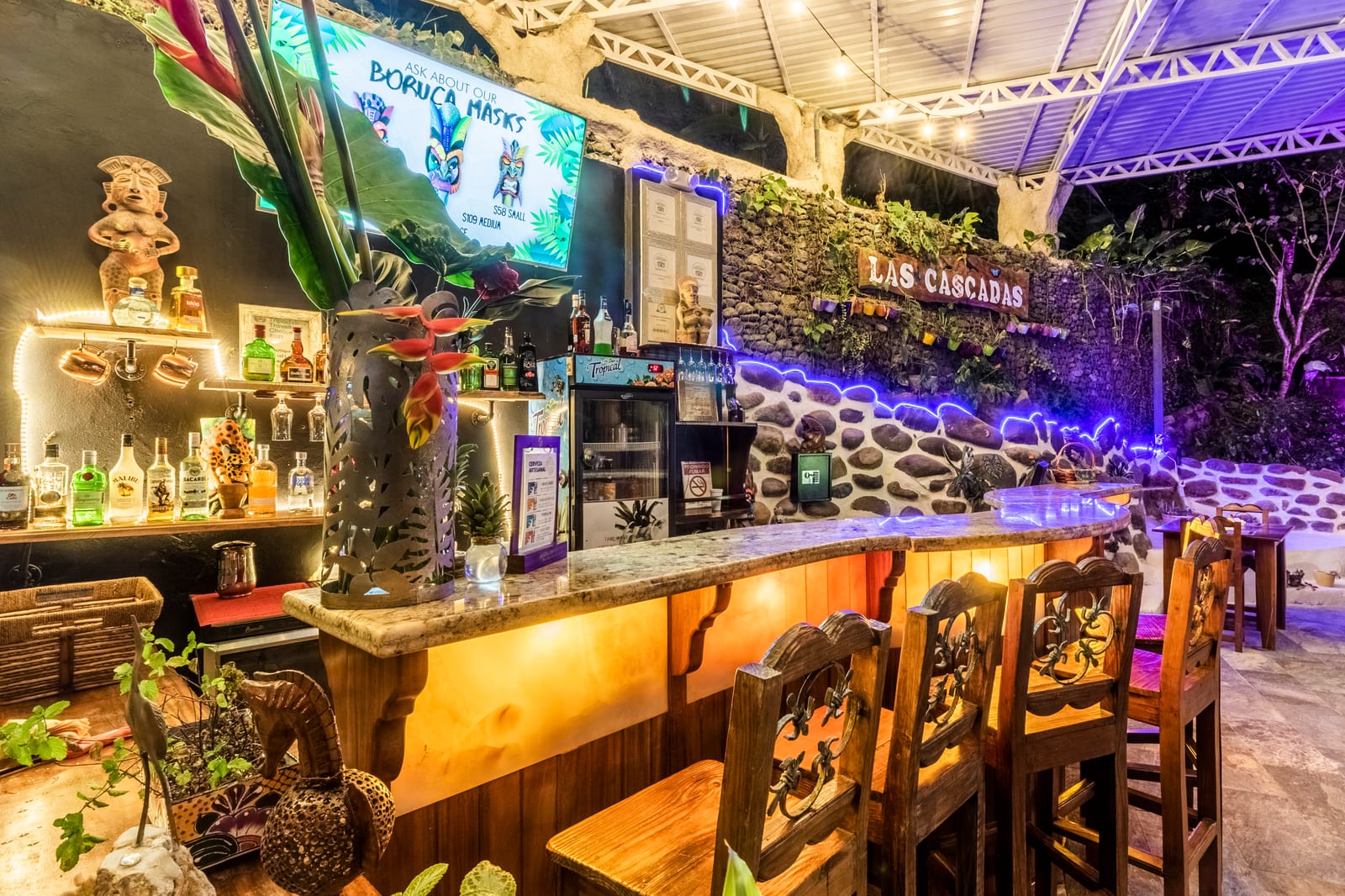 The bar counter close up, with carved wooden stools, bottles ranked on the back bar and a carved figure at the corner