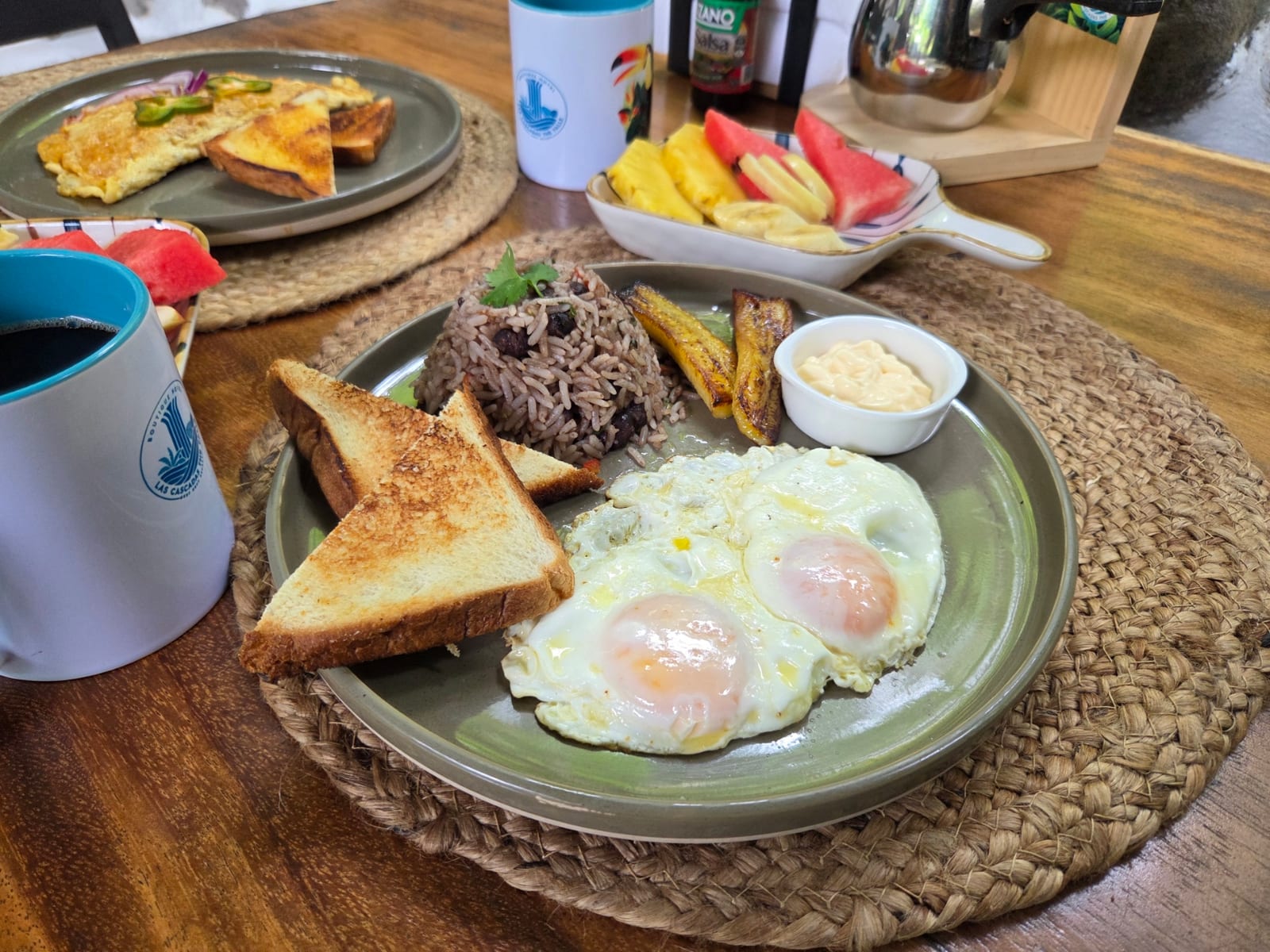 A breakfast plate of rice and beans, two fried eggs, toast and fried sweet plantain with a pot of sour cream, on a woven mat beside a bowl of pineapple, watermelon and banana and a coffee in a mug carrying the hotel's mark