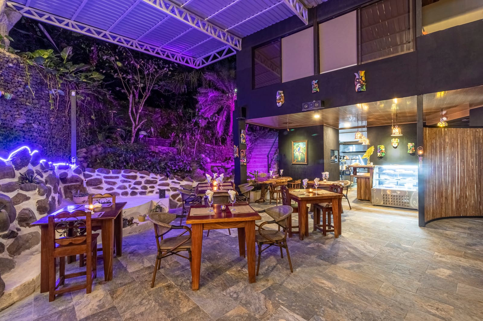 The covered end of the restaurant at night, with painted Boruca masks on dark walls, pierced ceramic pendant lamps and a lit glass dessert case beside the kitchen