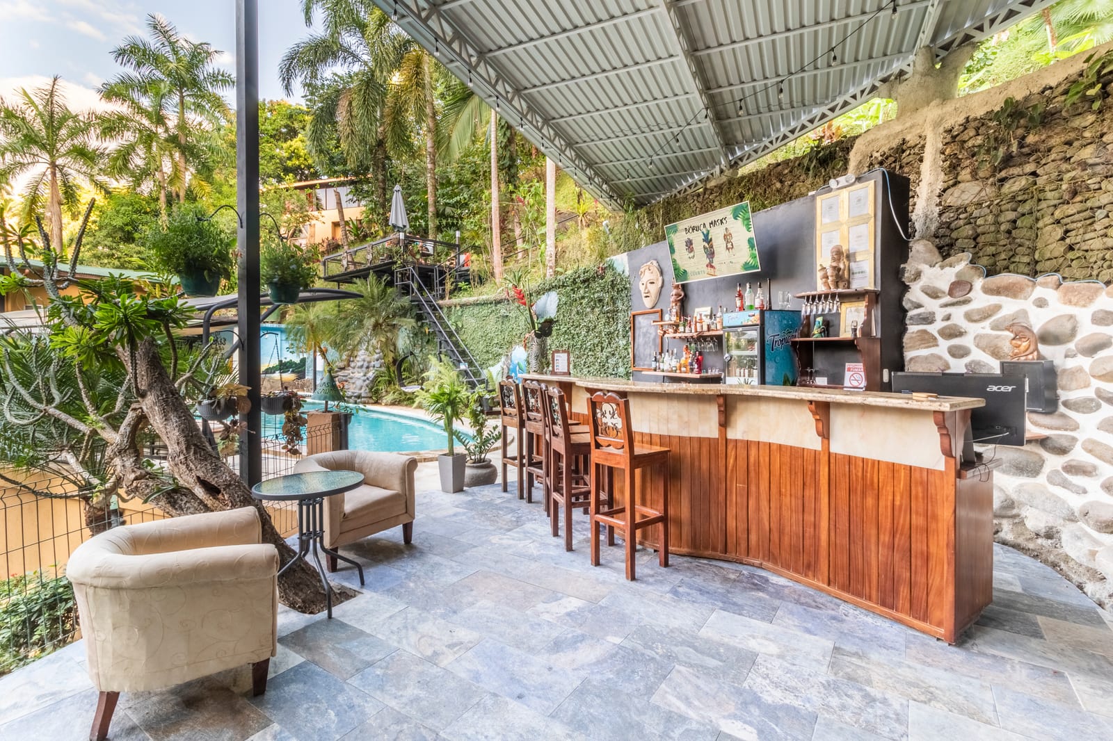The full run of the poolside bar with its stools, the pool and its painted wall behind, palms and the hillside beyond