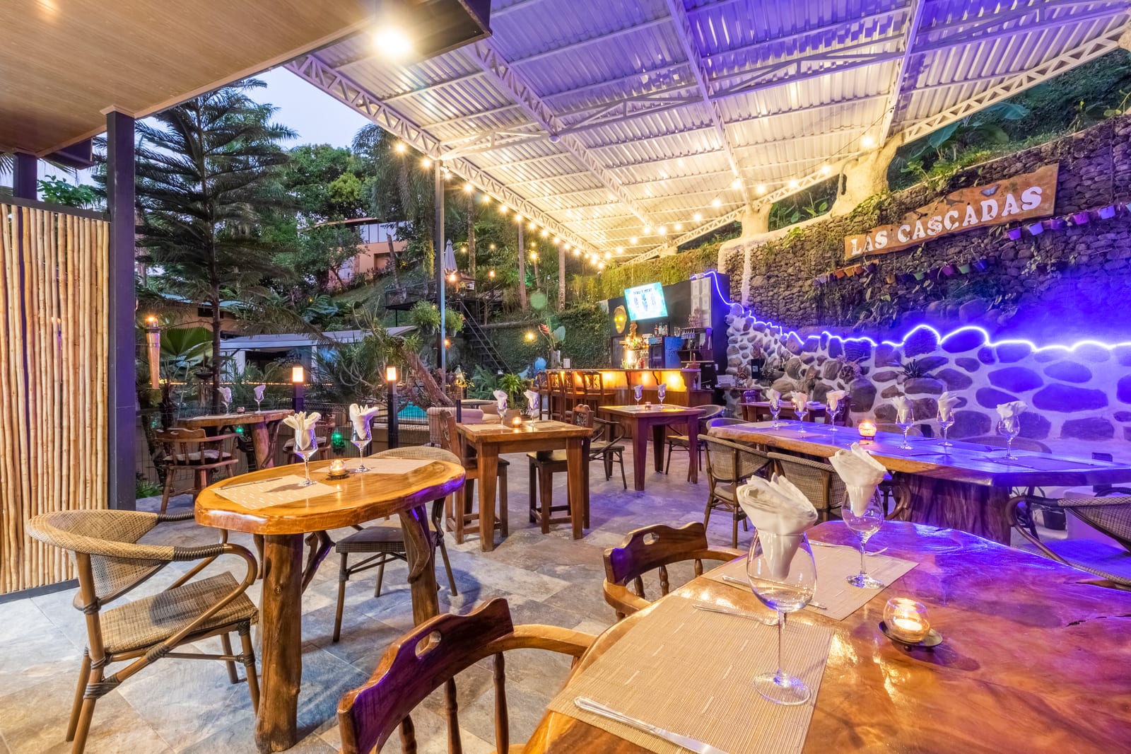 Wooden tables set with wine glasses and folded napkins, candles lit, the stone wall behind washed in violet light