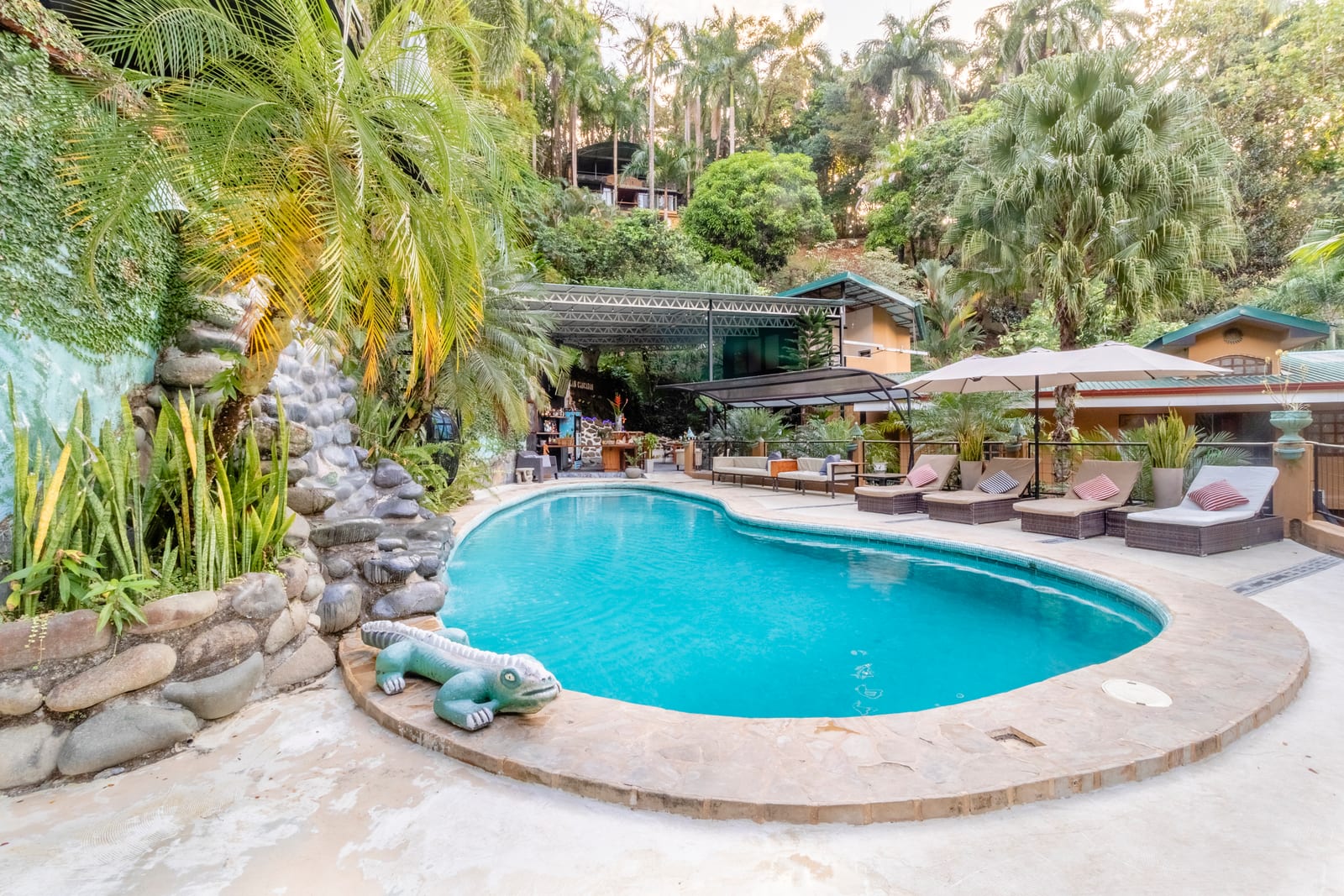 The man made waterfall running over river stones into the pool, with a carved stone iguana at the edge and the lit bar visible beyond