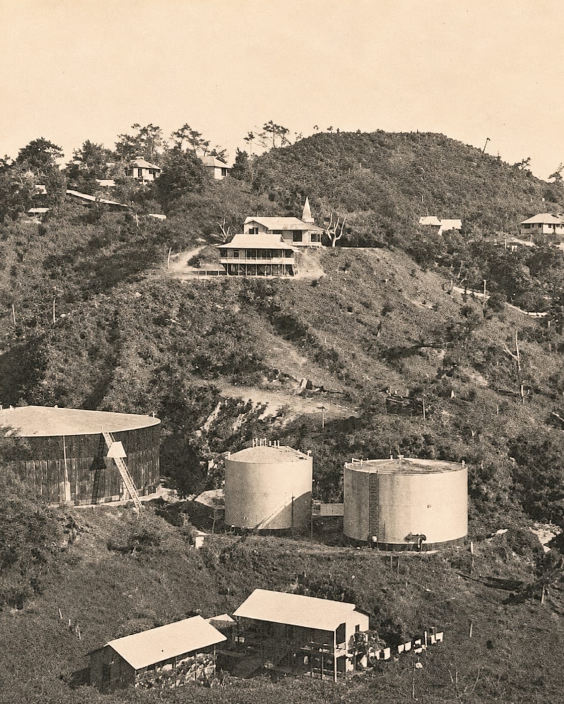 A sepia photograph of two large cylindrical storage tanks cut into a hillside, with wooden houses on the ridge above them and dense vegetation all around