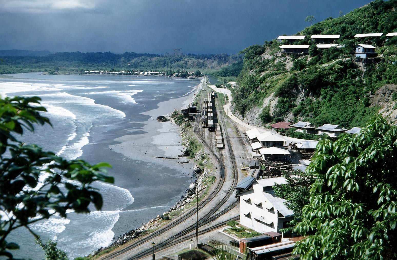 A colour photograph looking down a green hillside to surf breaking along a curving beach, with a road, a railway line and low buildings running between them