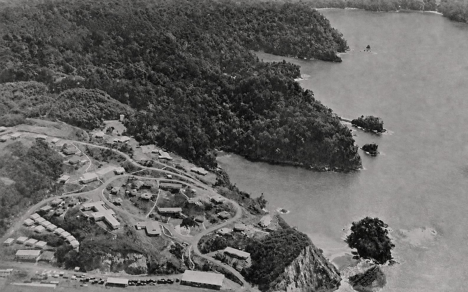 A black and white aerial photograph of a forested headland reaching into calm water, with a settlement of small buildings gathered along the shore at its base