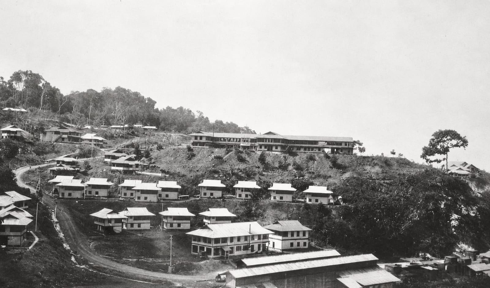 A black and white photograph of a hillside settlement, rows of small houses below a long two storey building on the ridge, with forest rising behind