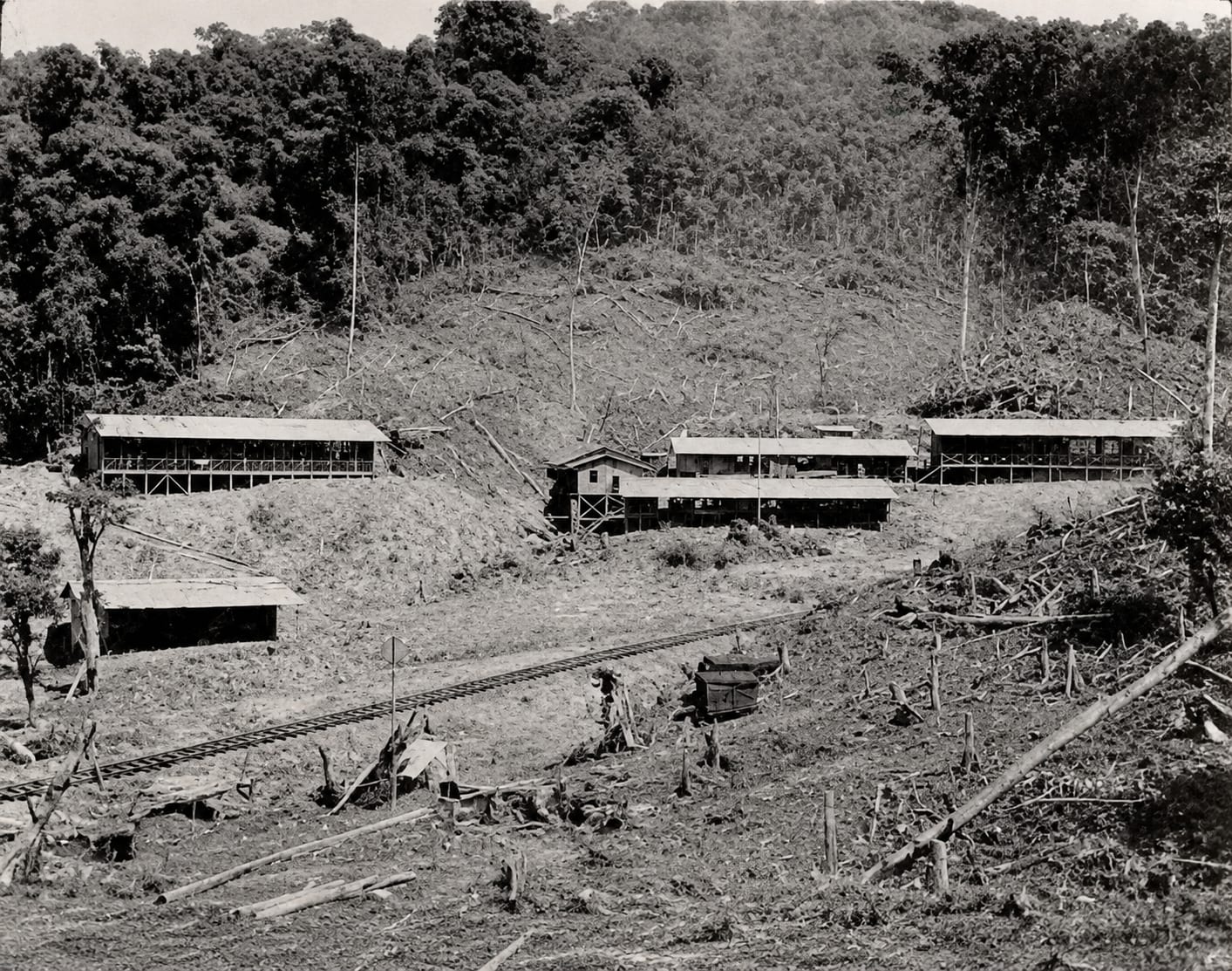 A black and white photograph of railway track running through newly cleared ground, with long timber sheds on the slope behind it and figures working near the line