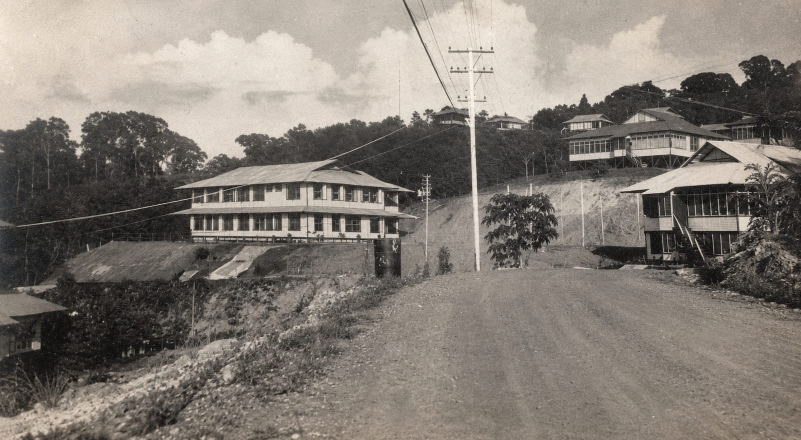 A sepia photograph of a wide dirt street running between two large two storey wooden buildings, with power lines overhead and more houses on the hill beyond