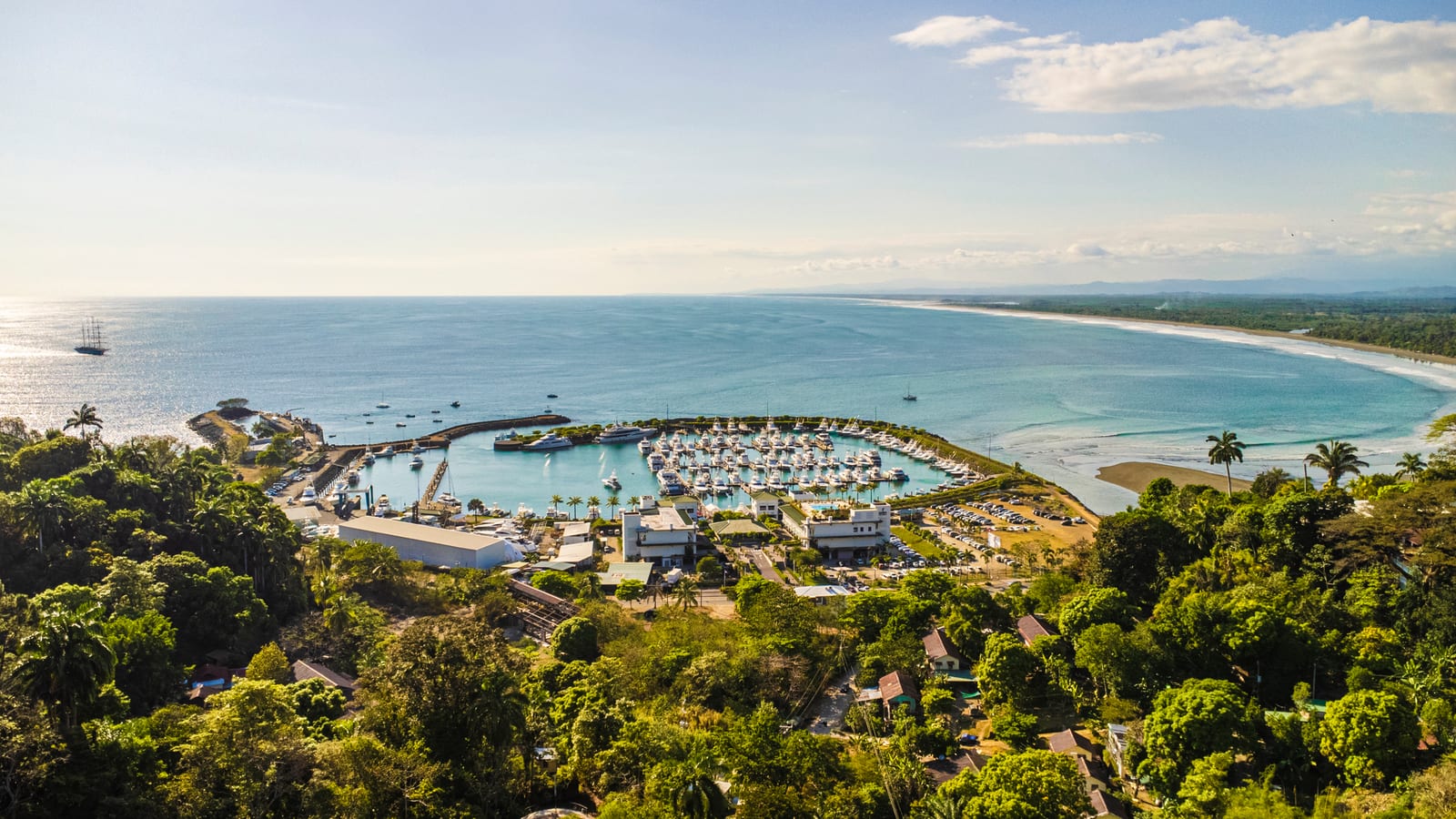 Marina Pez Vela from above, its docks full of sportfishing boats behind a curved breakwater, with the beach and the coastline running north beyond it