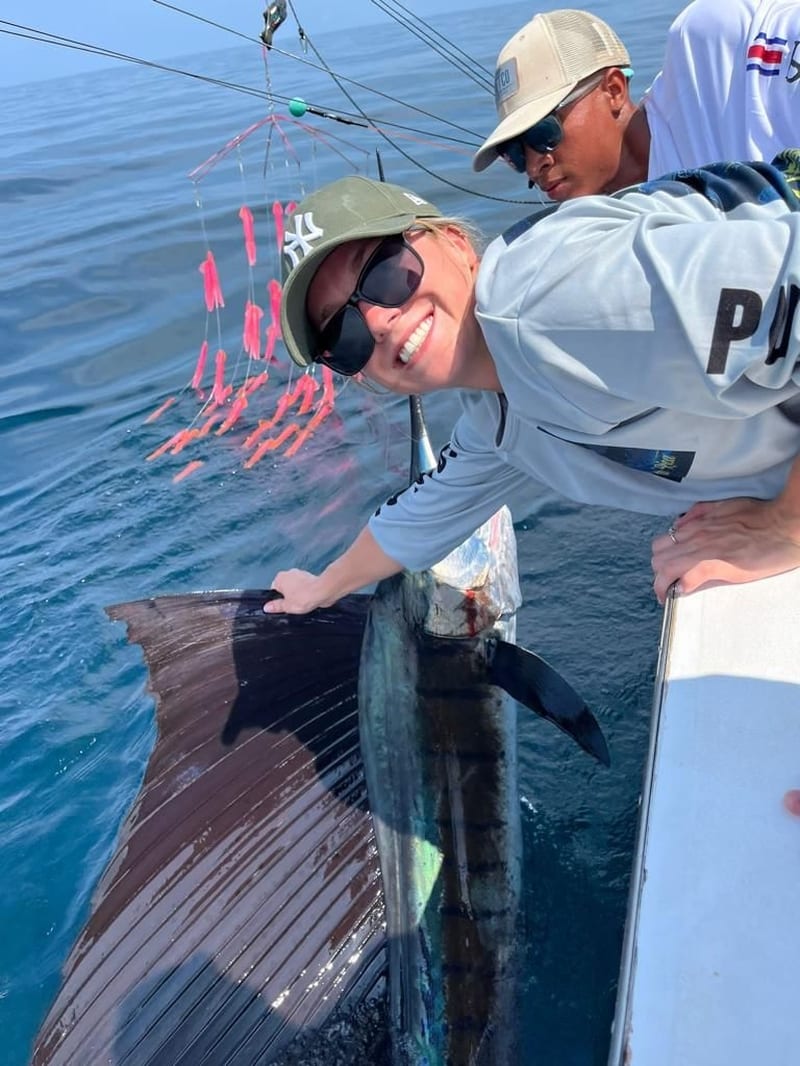 Un pescador sonriendo junto a un pez vela que un tripulante sostiene al costado del barco, con un aparejo de señuelos rosados colgando sobre el agua