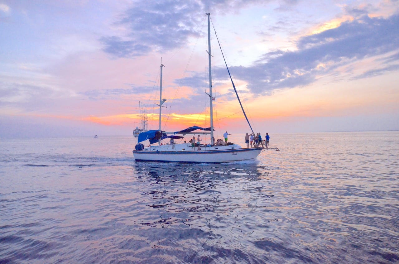 Un velero con gente de pie en cubierta, navegando en agua calmada al atardecer bajo un cielo naranja y violeta