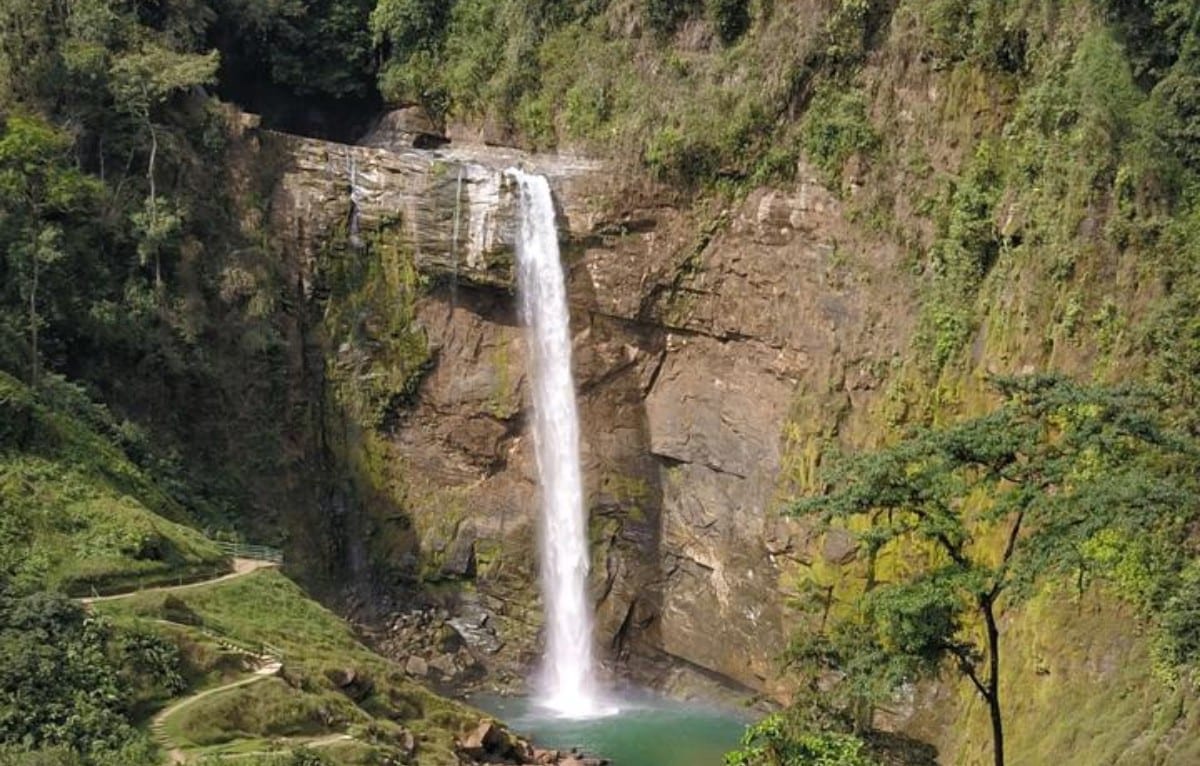 Una catarata alta cayendo por una pared de roca hacia una poza verde, con selva a los lados y un sendero por la orilla cercana