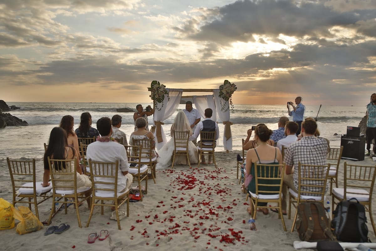 Una ceremonia en la arena al atardecer, con invitados sentados en sillas de madera a ambos lados de un pasillo cubierto de pétalos rojos, frente a un arco de flores a la orilla del mar