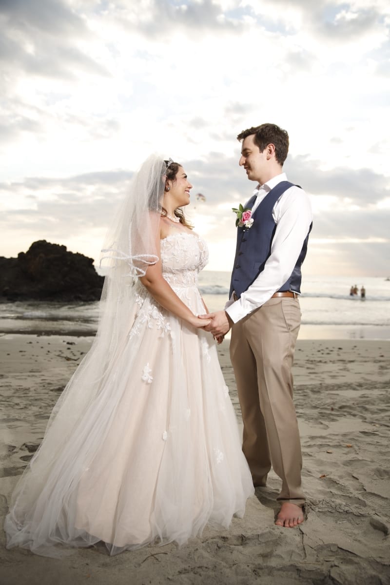Una novia de vestido blanco largo y velo frente a un novio de chaleco azul marino, tomados de la mano sobre la arena mojada al anochecer, con el mar y una roca oscura detrás