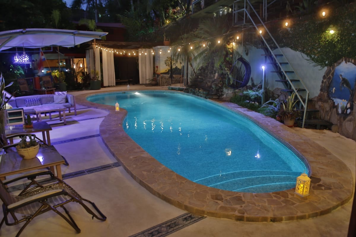 La piscina iluminada de turquesa por la noche bajo hileras de luces cálidas, con sillones y un bar iluminado a un lado y gradas pintadas que suben al fondo
