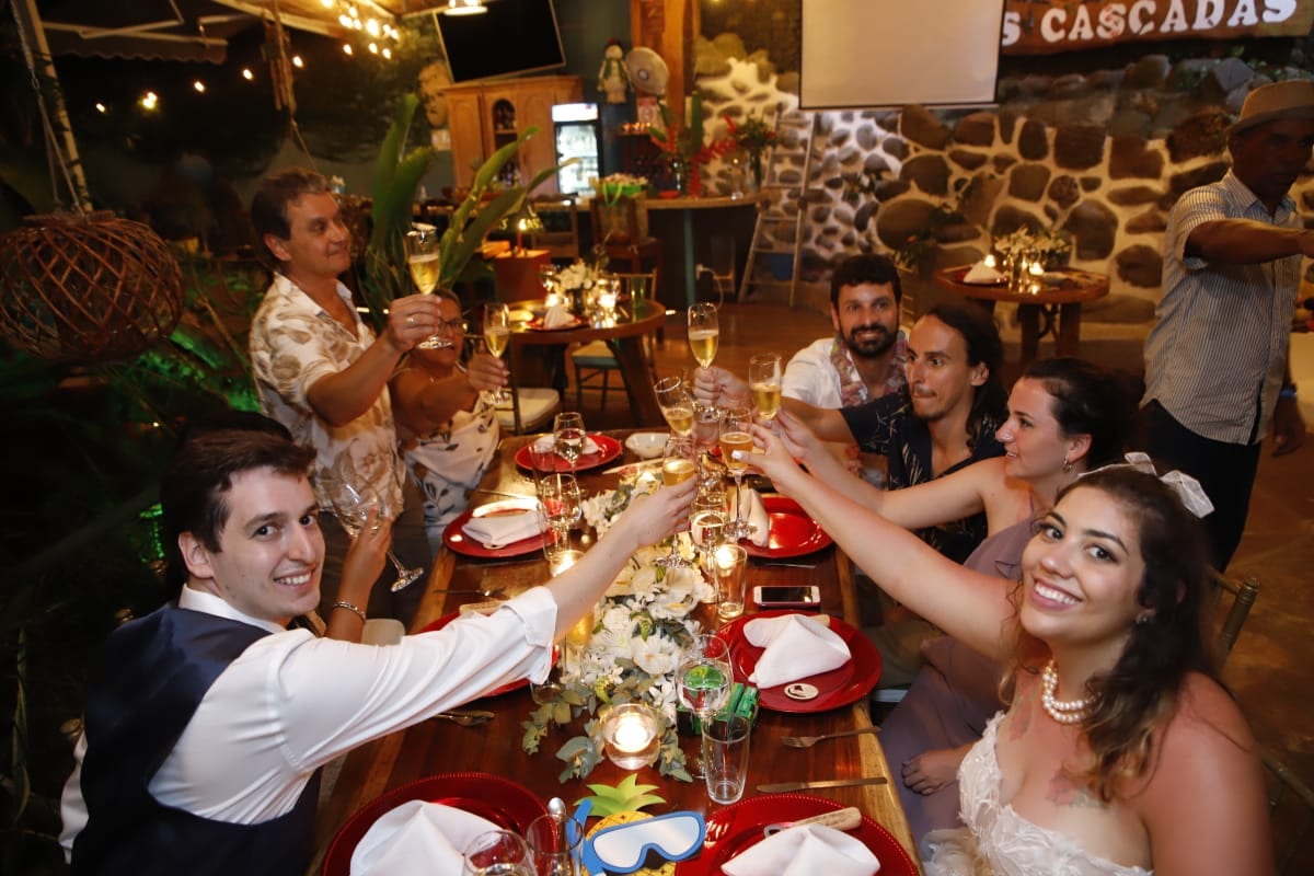 Invitados de una boda alrededor de una mesa larga de madera con luces colgadas, levantando copas de champán hacia la novia, con un rótulo de Las Cascadas en la pared de piedra de río detrás