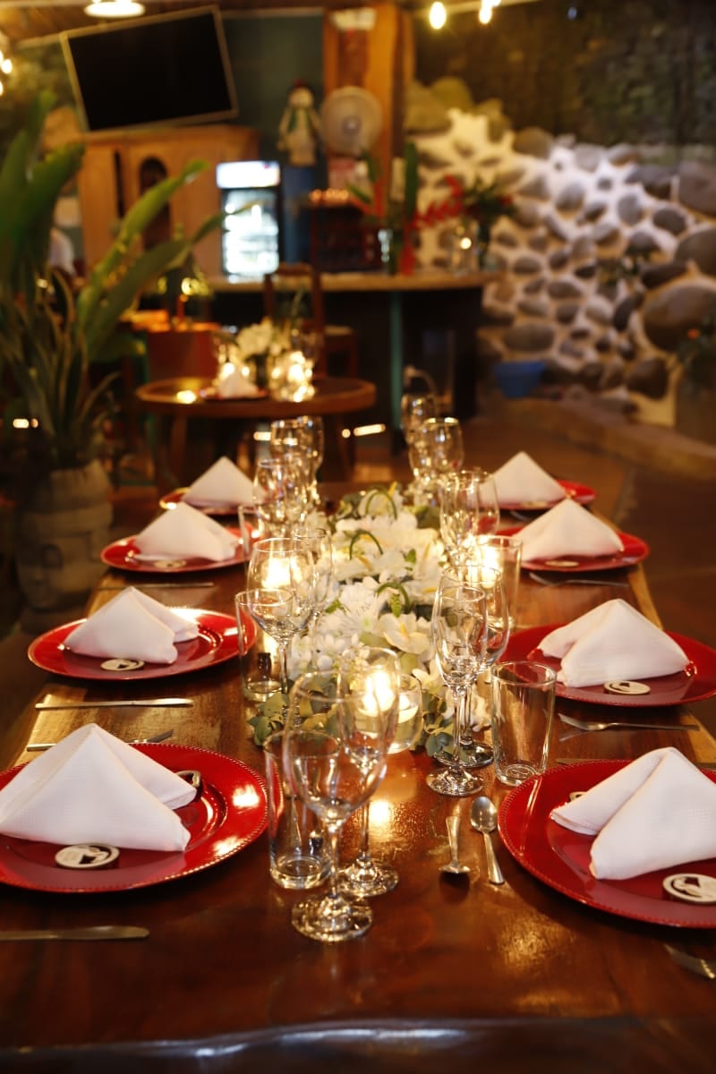 Una mesa larga de madera de canto natural puesta para una cena de boda, con bajoplatos rojos y servilletas blancas dobladas, hileras de copas de vino y una línea de candelas por el centro, contra una pared iluminada de piedra de río